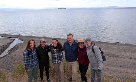 Six people stand, arms over one another, along a shoreline. A lake is in the background.