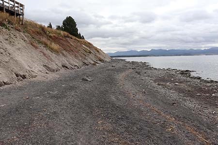 View looking down a tan and grey sand shoreline. In the left of a frame is the coastal bluff and to the right is a lake.