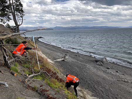 Two people wearing orange vests take measurement along a coastal bluff that is covered in light vegetation and debris. A lake is in the backround.