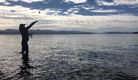 Silhouette of a person standing in a lake throwing a Sonobuoy into the water. The Sounobuoy looks like a small ball with a string attached.