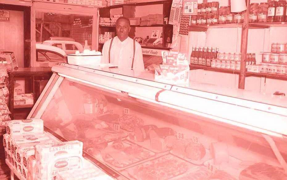 A red-tinted photo of a man standing behind a deli counter in a small grocery store