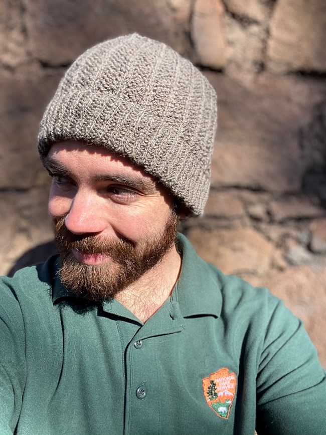 A person with a beard wearing a grey knit hat and a green polo shirt with the NPS arrowhead on it
