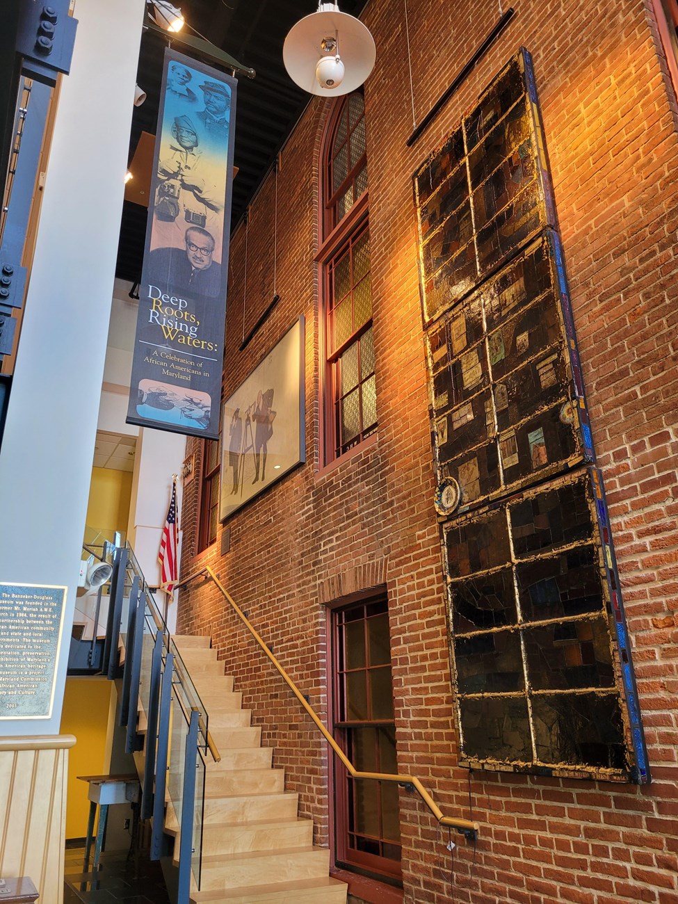 Photograph of brick building inside a more modern building. Stairs transcend underneath a banner with text that reads "Deep Roots, Rising Waters."