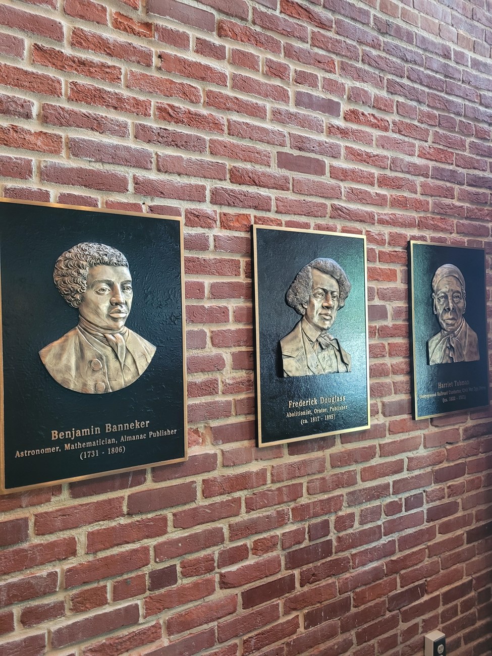 Photograph of three plaques fixed to a brick wall. From left to right, the plaques depict Benjamin Banneker, Frederick Douglass, and Harriet Tubman
