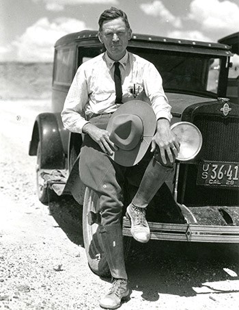 Frank Pinkly sitting on an old car with a foot on the fender and a wide-brimmed hat in his hand.