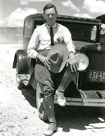 img014 A man wearing breeches sits on the front of his car with his left foot propped up against it. He holds his hat in his right hand.