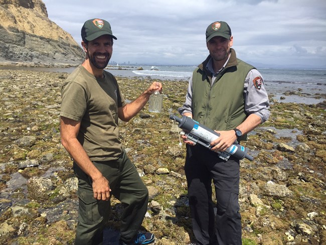 Pair of scientists stand on a rocky coast below towering bluffs. One holds a freshly collected water sample in a glass bottle as the other holds a large, cylindrical water sample collection device.