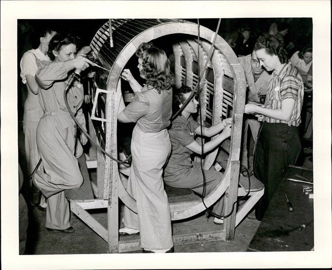 Industry Training in Wichita Black and white photo of five white women in pants or coveralls screwing bolts onto various parts of a cylindrical frame