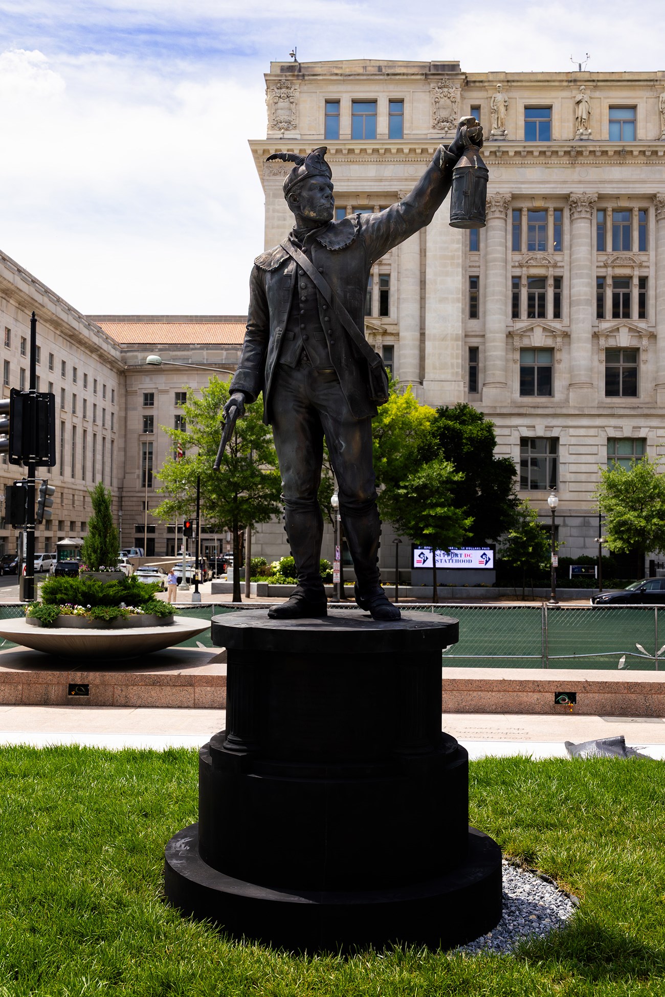 Dark bronze statue of a man in a feathered cap holding a lantern and a pistol