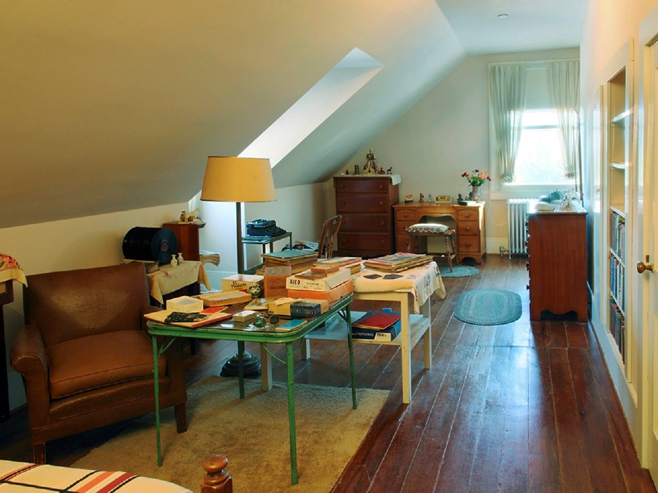 A loft bedroom with several dressers, twin bed and tables filled with books and papers. A typewriter sits at a table in front of a window.