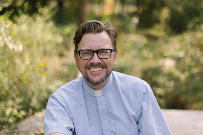 Man wearing glasses and a light blue shirt with a clerical collar smiles at camera.