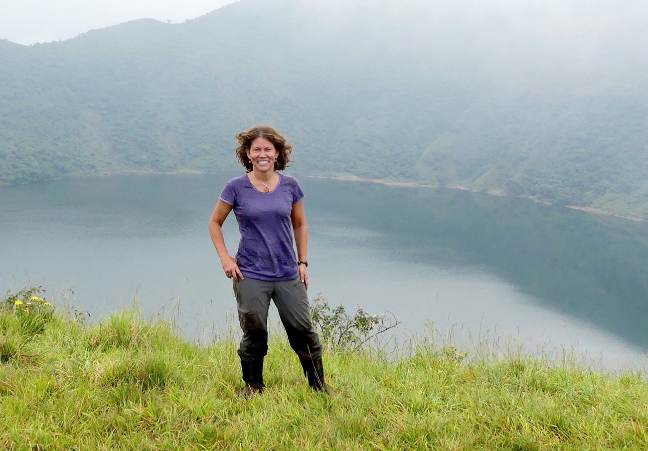 Dr. Jena Hickey Jena standing in the grass, with peaks and a crater lake behind her.