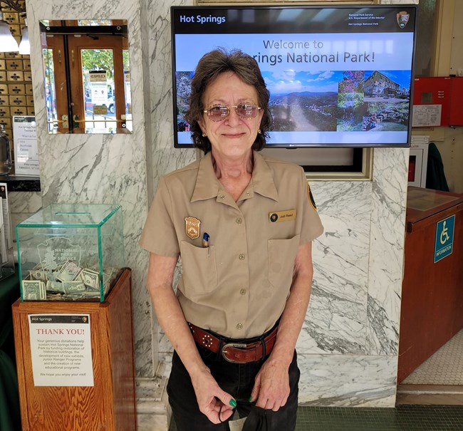 A middle-aged white woman with short brown hair and glasses poses in a tan NPS volunteer uniform.