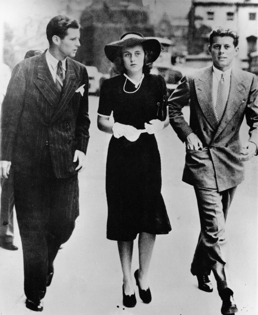 A black and white photo of a young woman in a hat, dress and gloves walking between two young men in suits on a city street. The man at left speaks, turned toward the woman, who looks ahead nervously. The man at right faces the viewer.