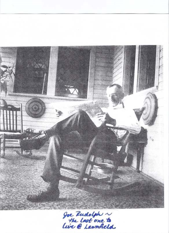 man with glasses sitting in a rocking chair on a porch while reading the paper