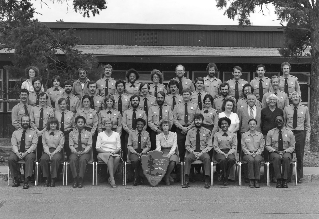 NPS Ranger Skills Class, Spring 1982 NPS employees pose outdoors in four rows for a class photo. Most wear NPS uniforms but a few have on street clothes.