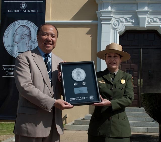 NPS Quarter Event Josie Fernandez in uniform stands with a man in a suit and tie. They each hold onto the sides of a framed NPS quarter set with drawing.