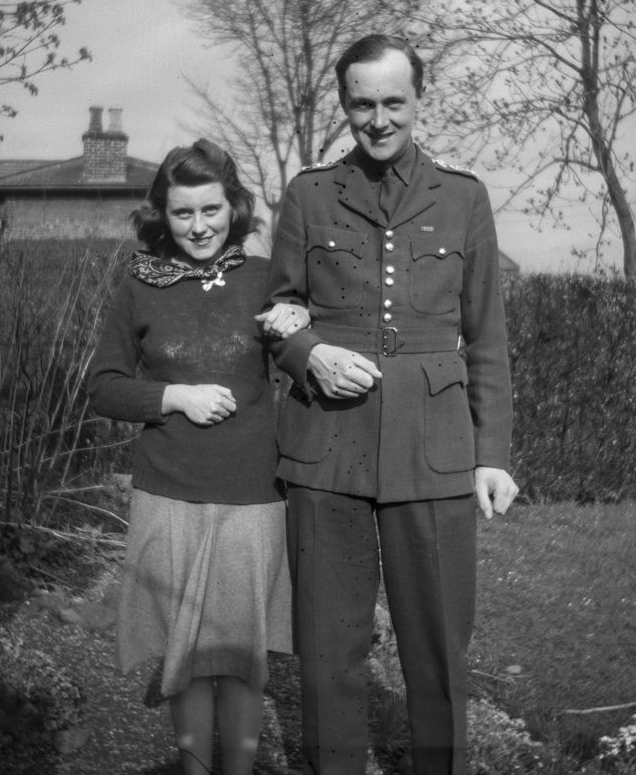 A black and white photo of a young woman and tall young man standing arm in arm on a lawn. She wears a silk necktie, sweater, and skirt, and he wears a khaki service dress military uniform. Behind them are hedges, trees, and a brick cottage.