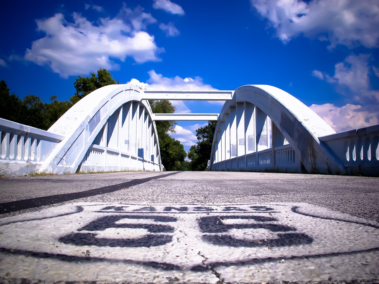 A road with a white emblem that reads "Kansas Route 66" leading up to a white truss bridge. A road with a white emblem that reads "Kansas Route 66" leading up to a white truss bridge.