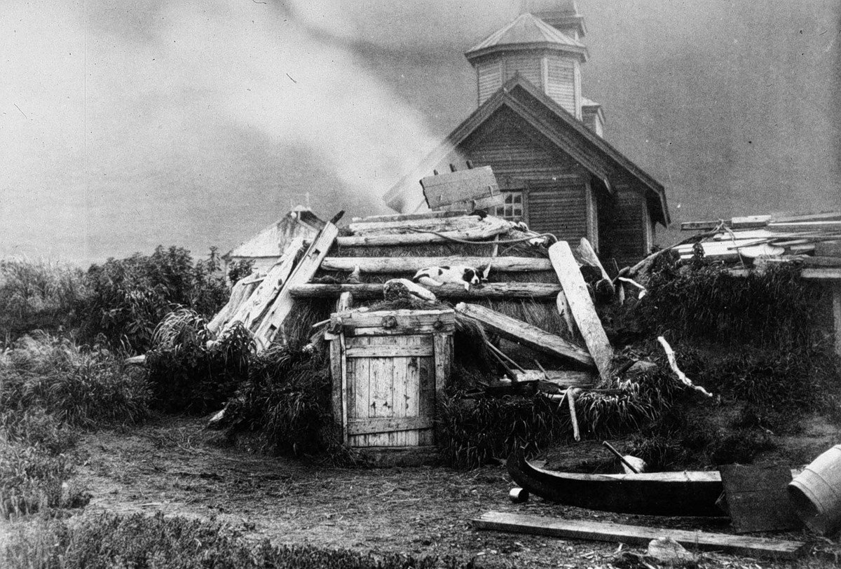 Historic photo showing a traditional Alutiiq sod house in the foreground. The Ascension of Our Lord Church is in the background.