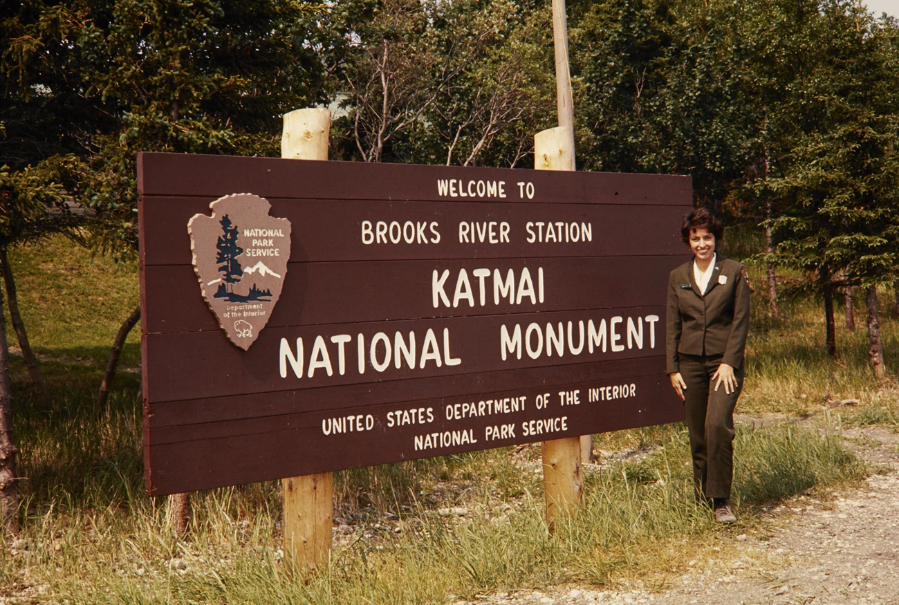 1969 Arrowhead plaque Woman in NPS uniform stands in front of Brooks River sign at Katmai
