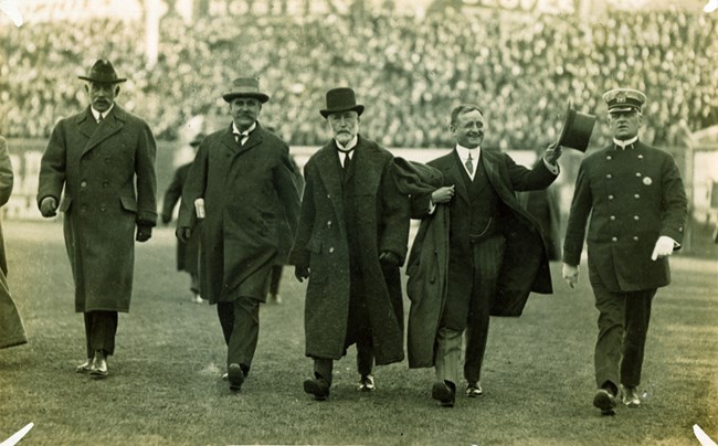 A sepia toned photograph taken on the field at Fenway Park.  In the forefront are five men walking toward the camera.  The four men on the left are dressed in formal wear, the man on the right is a police officer.  A crowd can be seen in the stands behind