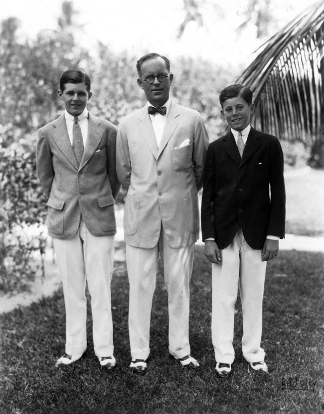 The three Kennedys are standing on grass in summer formal attire.  They stand in front of lush vegetation.