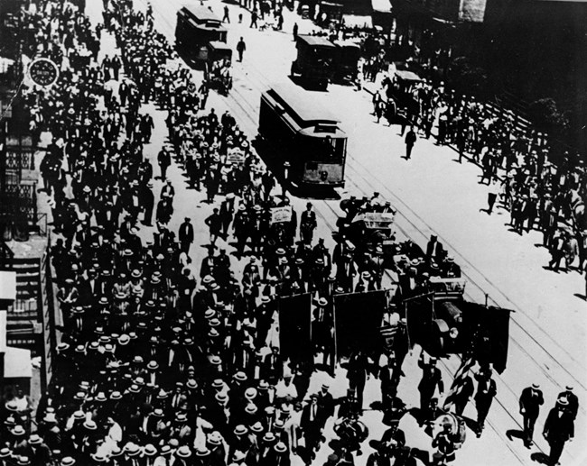 Marchers with banners supporting the 1910 Chicago Cloakmakers strike share the street with trolleys, automobiles, musicians and others. Overhead view of a crowd of people walking down a city street with banners, near a trolley car.