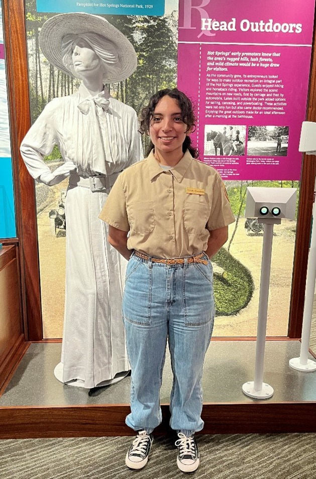 VIP Khristin A young woman with dark curly hair wearing a beige button-down shirt and jeans poses with her hands behind her back in front of a white state of a woman in a large hat.