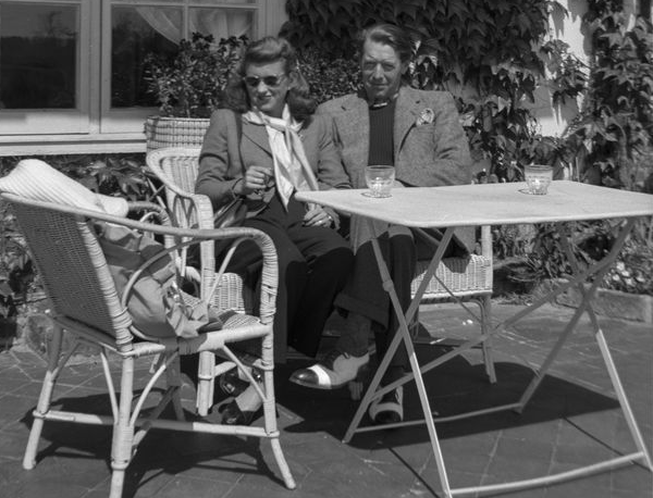 A black and white photo of a smiling young woman in a blazer, neck scarf, slacks, black flats and sunglasses sitting with a smiling man in a blazer, slacks and black toed white shoes at a white patio table, in front of a building covered in ivy leaves.