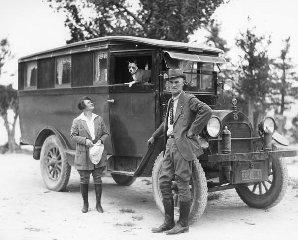A man and woman stand next to a large camping vehicle as a dog looks out the window
