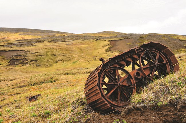 The Kiska Battlefield Today Color photo of grassy field, fog, and a rusted track assembly.