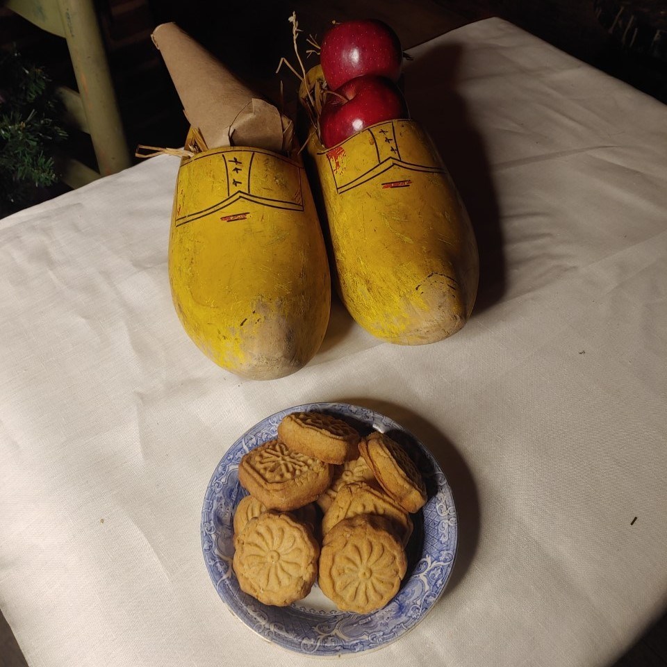 Four small round cookies sit on a plate. Next to them, large wooden shoes filled with apples.
