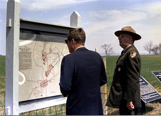 President Kennedy in a suit stands with a uniformed National Park Service ranger.  Kennedy is looking at a large map.