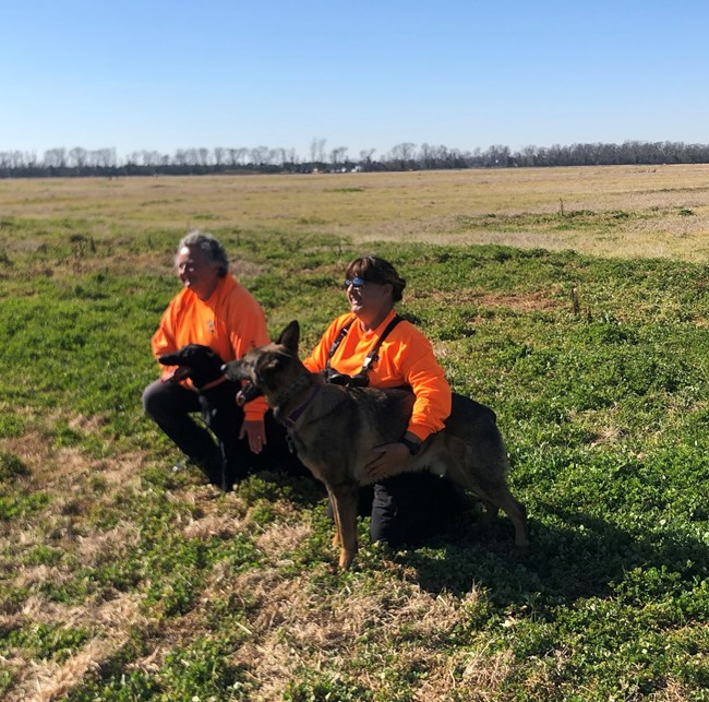 2 dogs and 2 people kneeling in a field.