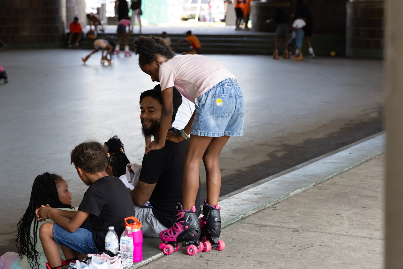 Lenny Webb smiles at his four young children A dad smiles at his four young children as they take a water break on the steps of the rink at Anacostia Park Skating Pavilion