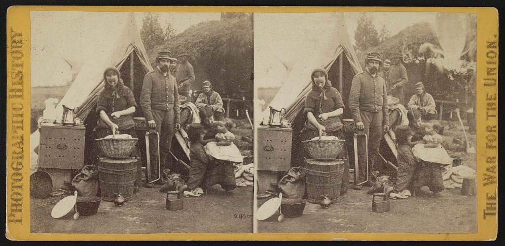A stereoscope image of a woman in an army camp holding a basket of laundry.
