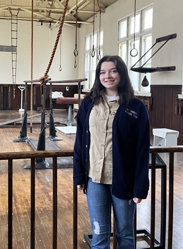 A young woman with dark straight hair wearing a navy-blue cardigan and jeans poses in front of a historic gymnasium with old, wooden equipment.
