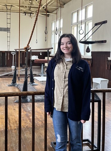 VIP Lily C. A young woman with dark straight hair wearing a navy-blue cardigan and jeans poses in front of a historic gymnasium with old, wooden equipment.