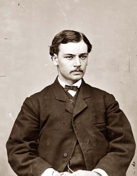 man in his early 20s sitting with his hands folded. He has a dark suit and mustache