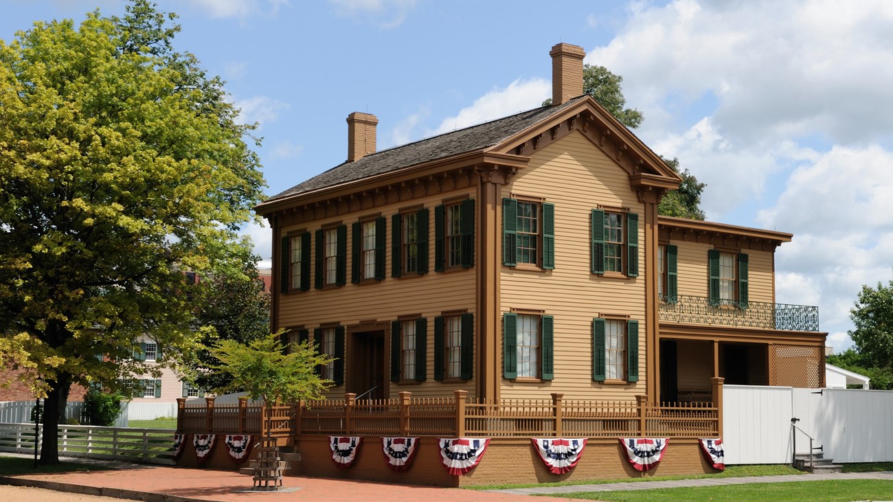 A light orange two story home containing many windows with green shutters sits along the side of a street.