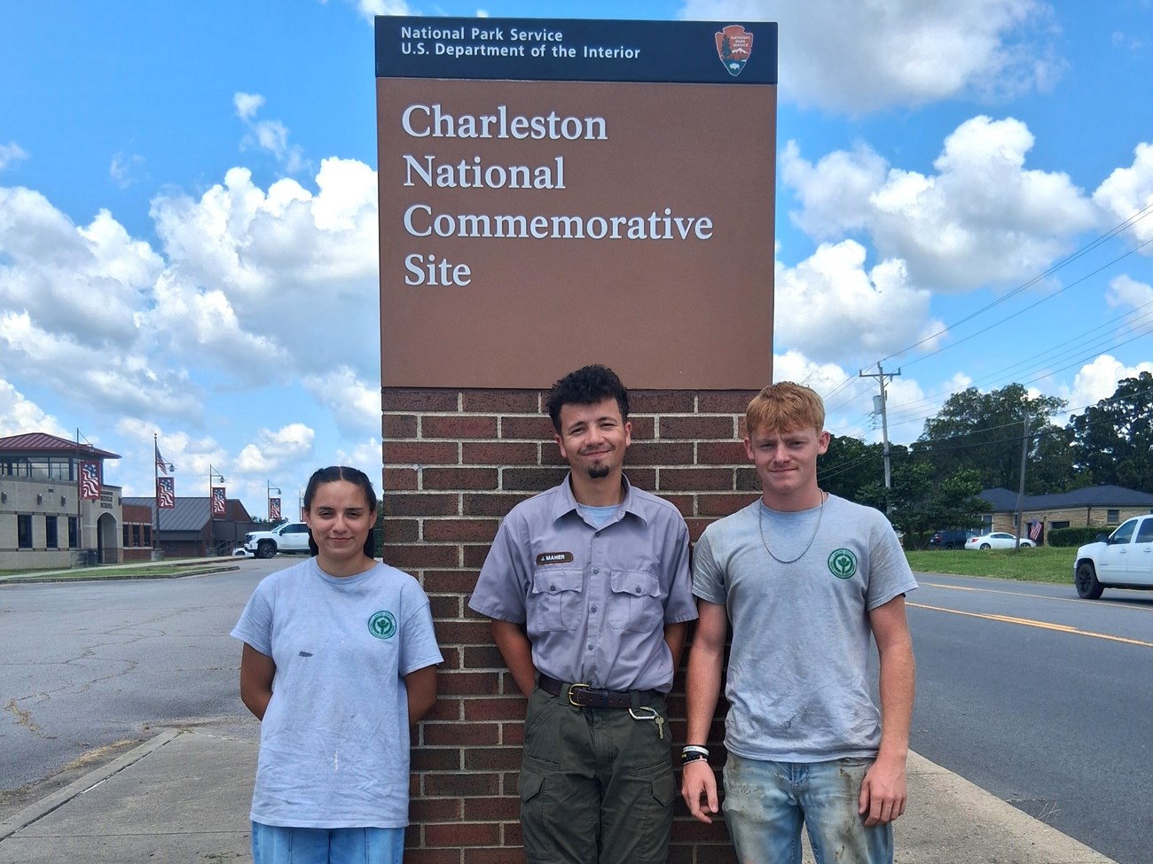 Three people stand side by side in front of a brick wall with a sign reading “National Park Service, U.S. Department of the Interior – Charleston National Commemorative Site.”