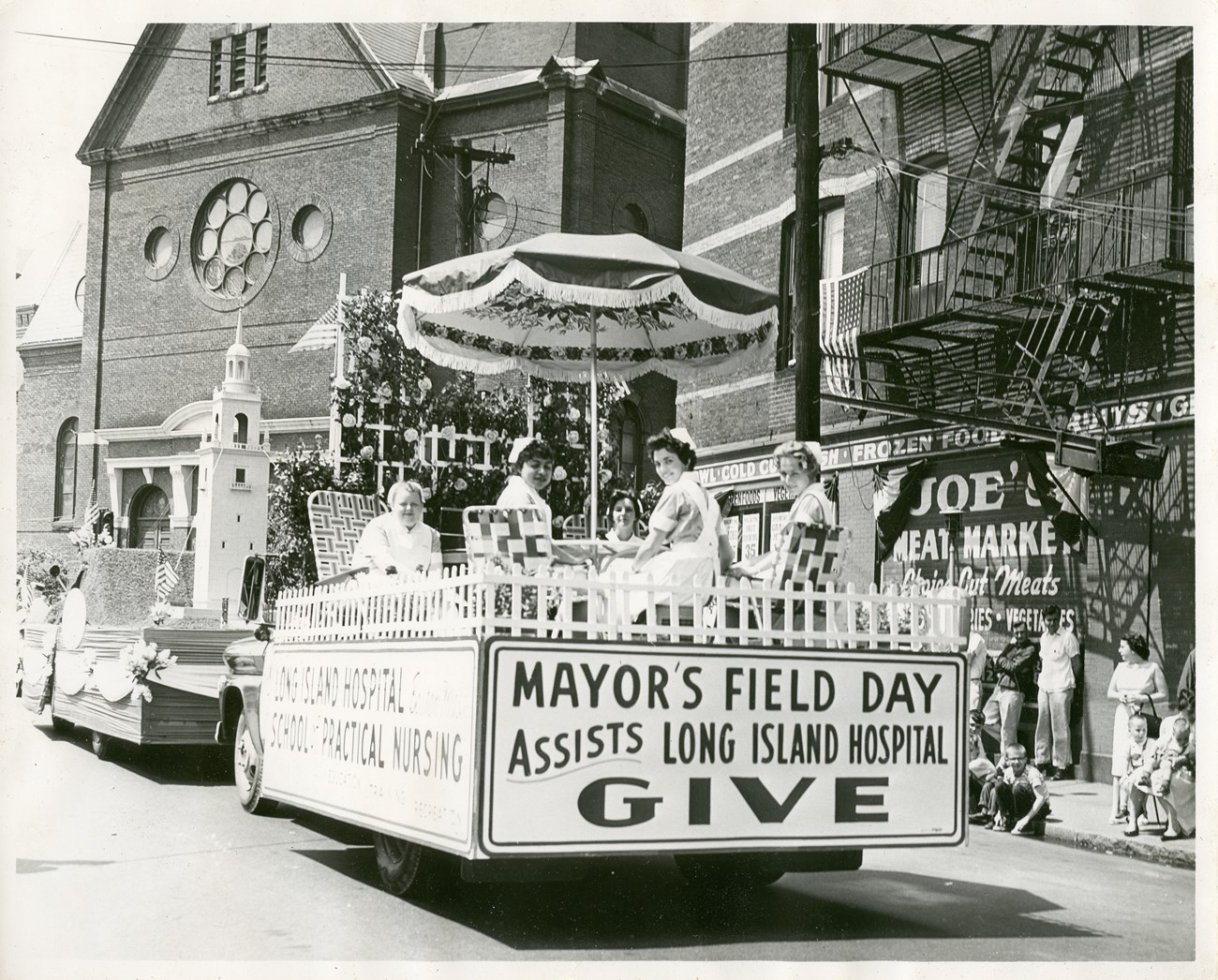 Five nurses sit on a parade float under an umbrella. Behind the float is another float with a miniature Dorchester Heights monument. Spectators view the float on the sidewalk to the right.