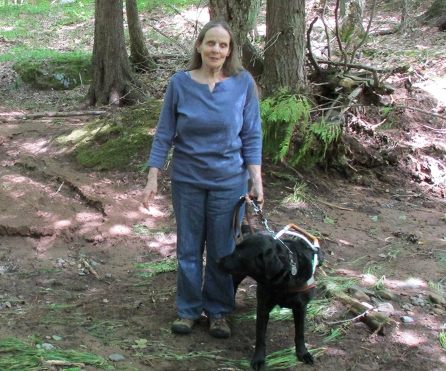 Lynda Boose and her guide dog Missy stand on a trail in a forest.