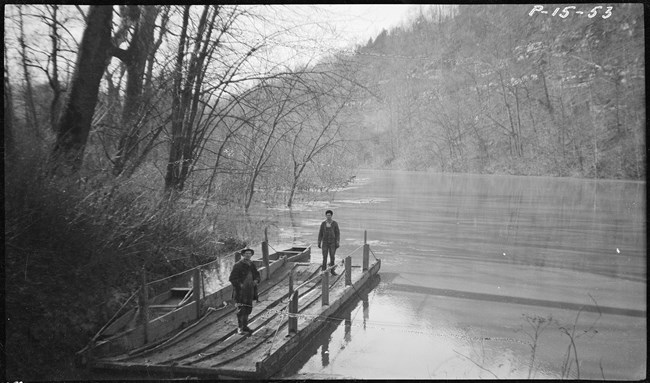 Two men stand on a wooden ferry boat floating on a wide river lined with trees.