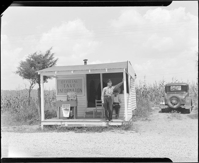 A black and white photo of a small road side shack with a man standing outside