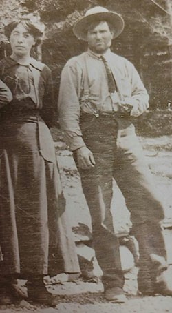 Mae and William Burns standing next to each other. She wears a dark long-sleeved shirt and ankle length skirt. He has on shirt, pants, tie, and hat.