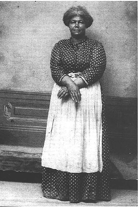 Mag Palm, showing how her hands were bound during her attempted abduction. A black and white photo of Mag Palm, showing how her hands were bound during her attempted abduction. She is wearing a dark dress with small white polka dots and a long white apron.