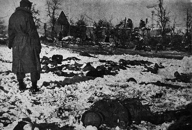 The Malmedy Massacre A black and white image showing a soldier looking over the bodies of fallen soldiers in the snow