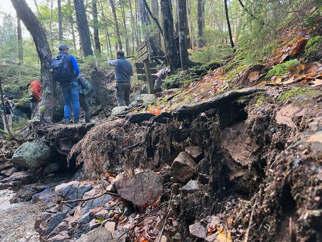 park staff stand on in tact section of trail with washed out trail, rocks, and roots exposed in front of them